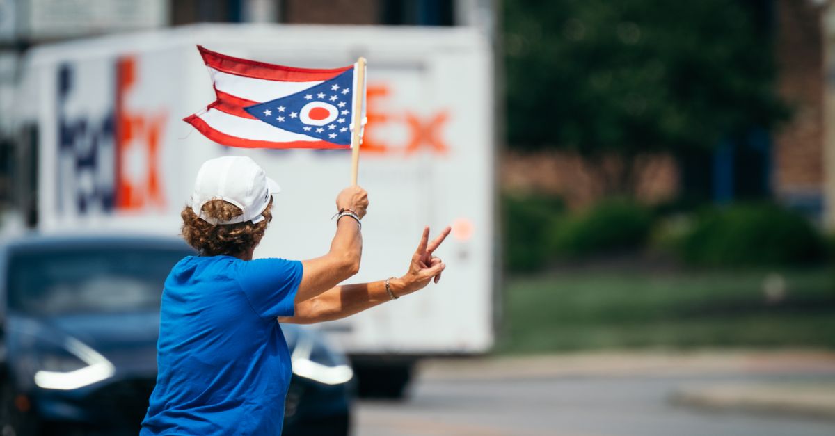 A person waves the Ohio state flag while making a peace sign on a city street.